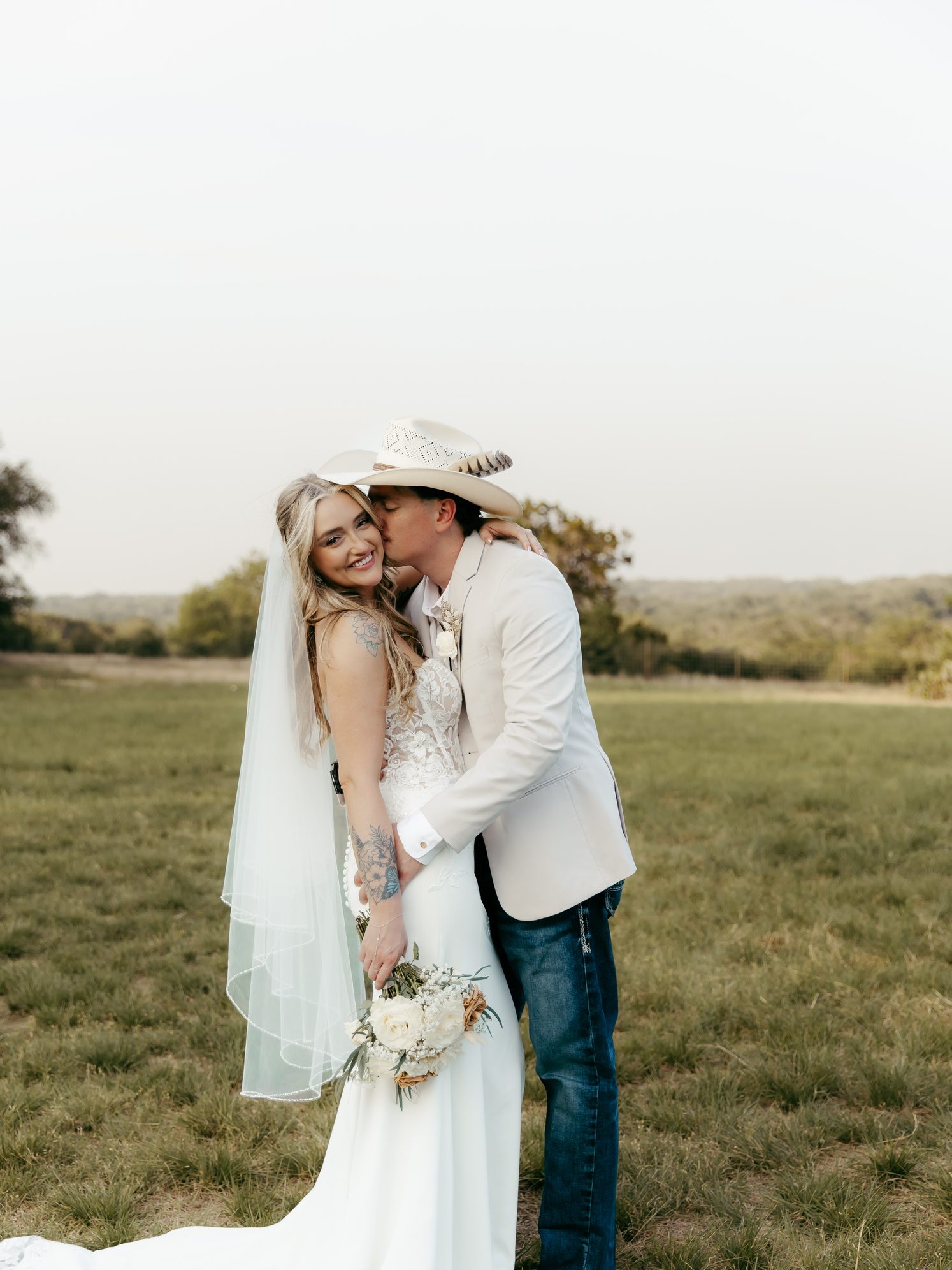 Ray and Cassidy in an open Hill Country field at golden hour, the groom in a cowboy hat kissing the bride's cheek