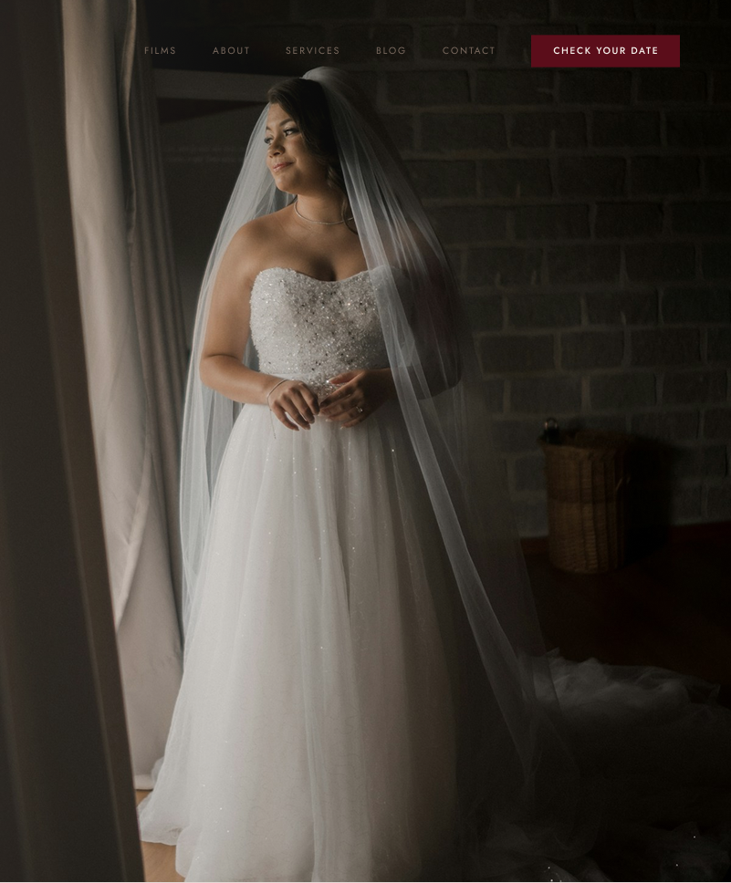 A bride in a full veil standing beside a window, lit by soft daylight against a brick wall